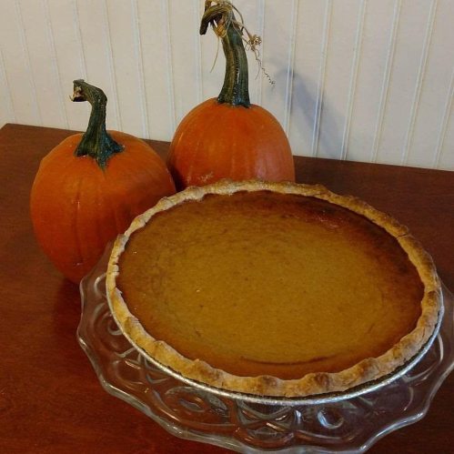 A pumpkin pie on a glass dish, with two decorative pumpkins in the background.