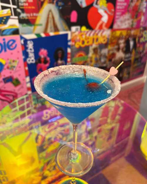 A vibrant blue cocktail in a glass with a sugared rim, garnished with a cherry and a heart-shaped pick, against a colorful backdrop.