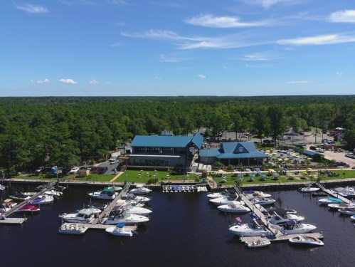 Aerial view of a marina with boats docked, surrounded by trees and a building with a blue roof. Clear blue sky above.