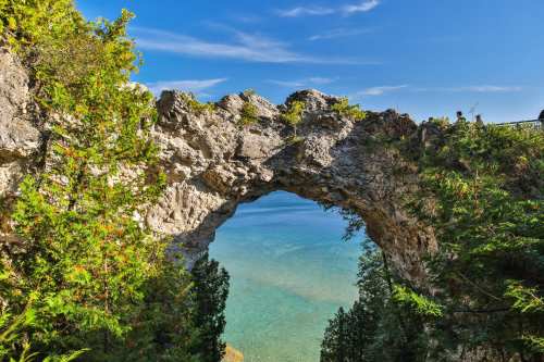 A natural stone arch frames a view of calm blue waters and lush greenery under a clear blue sky.