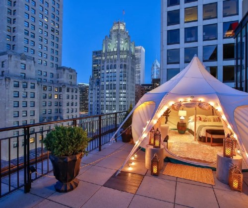 A cozy tent with lights on a rooftop terrace, overlooking a city skyline at dusk.