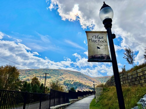A scenic pathway with a "Visit Flag Rock" banner, surrounded by mountains and a blue sky with clouds.