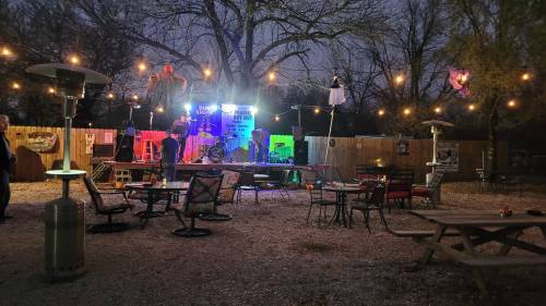 Outdoor stage with colorful lights, surrounded by seating and string lights, set in a cozy, gravel-filled venue.