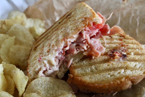 A grilled sandwich with ham and cheese, partially eaten, served with a side of potato chips on brown paper.