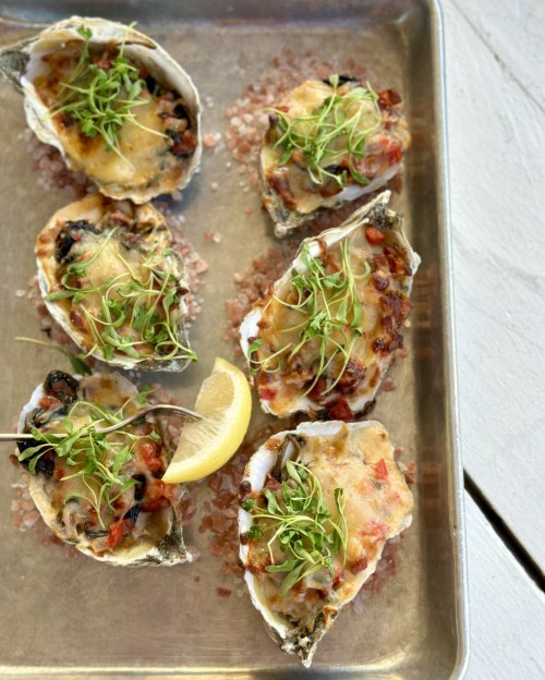 Baked oysters topped with cheese, herbs, and diced vegetables, served on a tray with a lemon wedge.