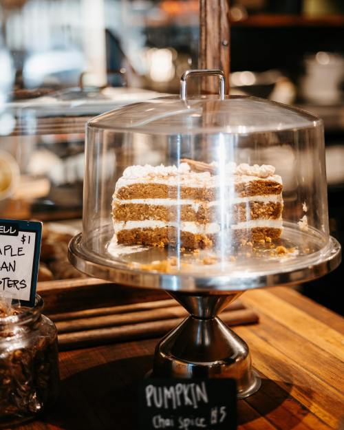 A layered pumpkin cake with cream frosting displayed under a glass dome on a wooden counter.