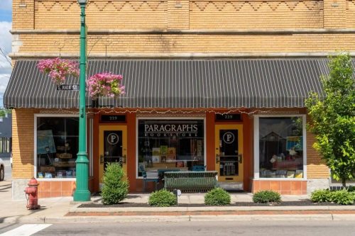 A charming bookstore with a striped awning, colorful flowers, and a green bench outside, set in a brick building.
