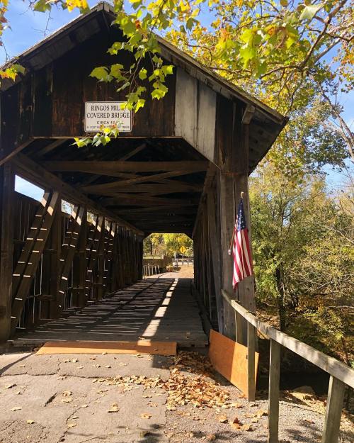 A rustic covered bridge with wooden beams, surrounded by autumn foliage and an American flag at the entrance.