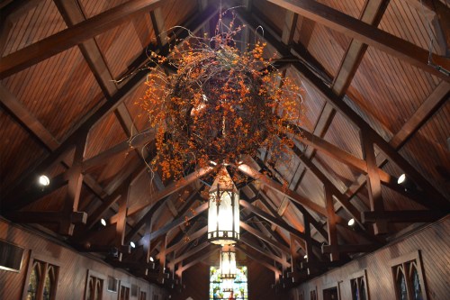 A wooden church interior with a high ceiling, featuring a large, orange floral arrangement hanging above.