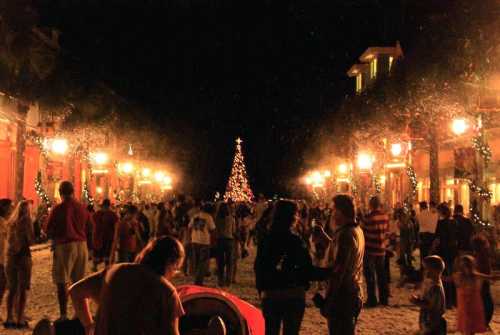 A festive street scene at night, bustling with people, decorated with lights and a Christmas tree in the background.