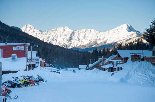 Snow-covered street in a mountain town, with snowmobiles parked and majestic peaks in the background.