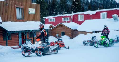 Snowmobiles parked in a snowy area, with riders preparing to depart near wooden cabins and a red building.