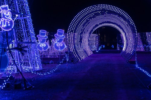 A pathway illuminated by vibrant blue and white lights, featuring snowman decorations and a glowing archway.