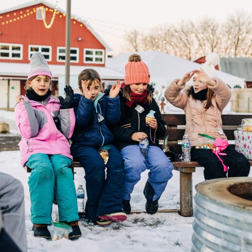 A group of children in winter clothing sitting on a bench, enjoying a snowy outdoor setting with festive decorations.