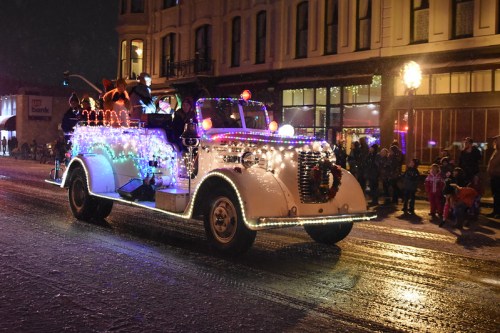 A festive parade float decorated with colorful lights, featuring people and a reindeer, driving down a street at night.