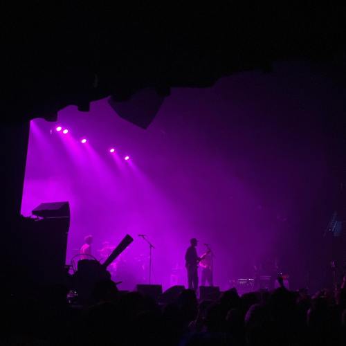 Silhouetted band performing on stage with purple lighting and a crowd in the foreground.