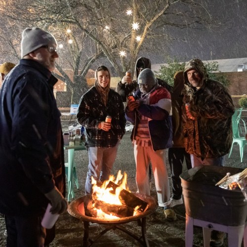 A group of people gathered around a fire pit, enjoying drinks and the snowy night atmosphere.