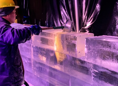 A worker in a hard hat uses a mallet to shape large blocks of ice, illuminated by purple lighting.
