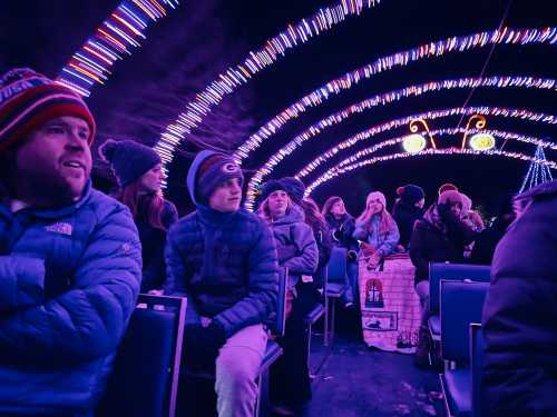 A group of people wearing winter clothing sits inside a brightly lit, festive tunnel of lights.