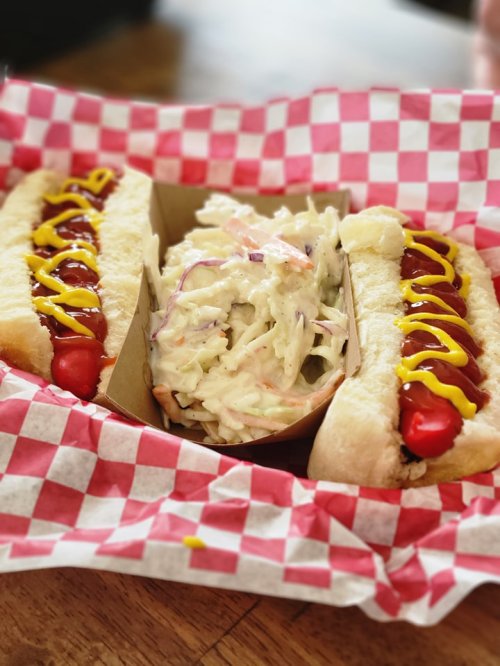 Two hot dogs with mustard and ketchup, served with a side of coleslaw in a red and white checkered basket.