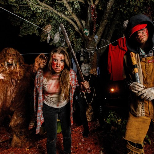 A group of people in horror-themed costumes, with eerie makeup and props, standing in a dark, outdoor setting.