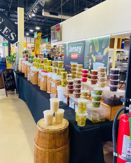 A display of various pickles in jars and cups at a market, with a sign for "Jersey Pickles" in the background.