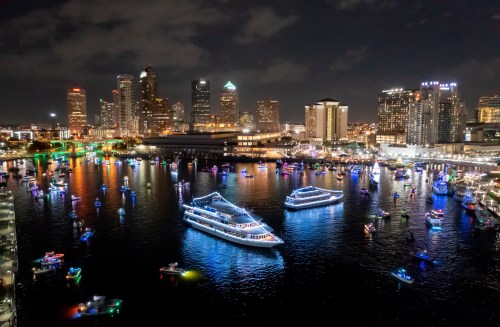 A vibrant city skyline at night with illuminated boats on the water, reflecting colorful lights.