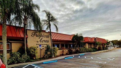 Exterior of Tropical Acres Steakhouse with palm trees and a cloudy sky, featuring a parking lot in front.