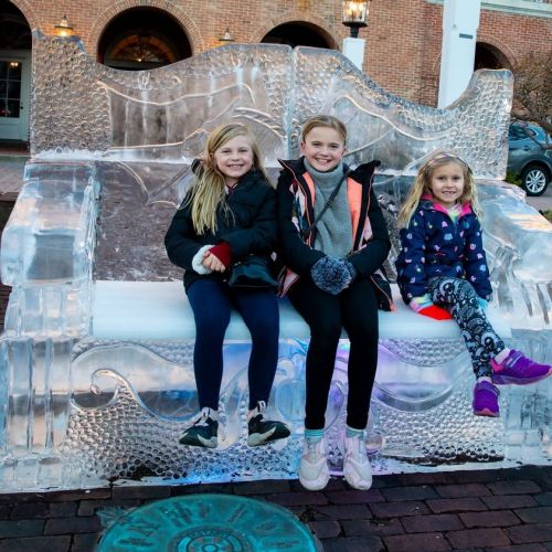 Three children sit on a large ice sculpture bench, smiling and bundled up in winter clothing.