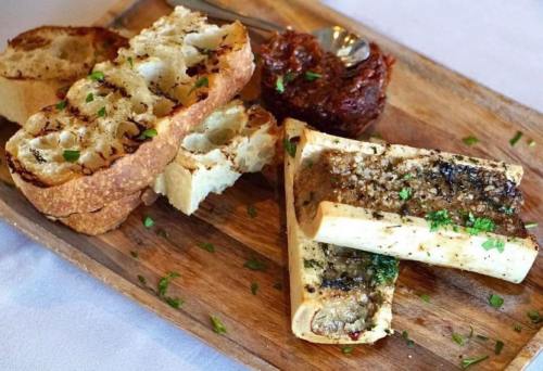 A wooden platter with grilled bread, roasted bone marrow, and a dollop of savory spread, garnished with herbs.