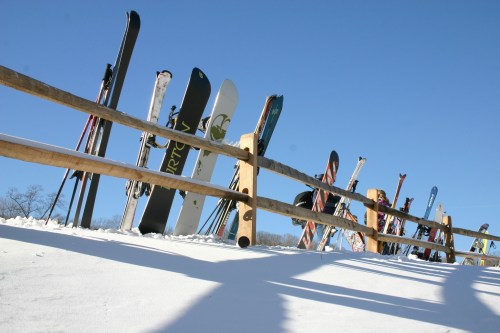 A row of skis leaning against a wooden fence, with a clear blue sky and snow-covered ground in the background.