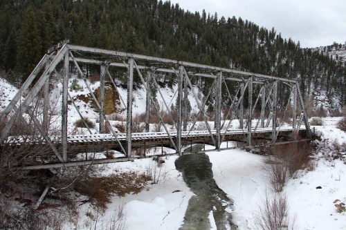 A weathered metal bridge spans a snowy landscape, with a river flowing beneath and mountains in the background.