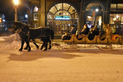 A horse-drawn sleigh carries passengers through a snowy street near a warmly lit restaurant.