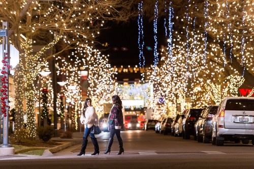 Two women walk across a festive street adorned with twinkling holiday lights and decorations at night.