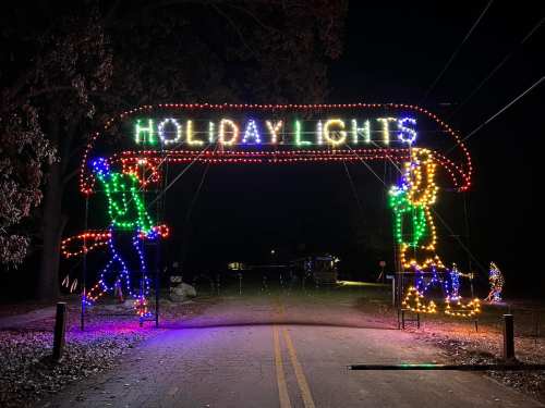 Colorful holiday lights arch over a road, featuring festive figures on either side, illuminating the night.