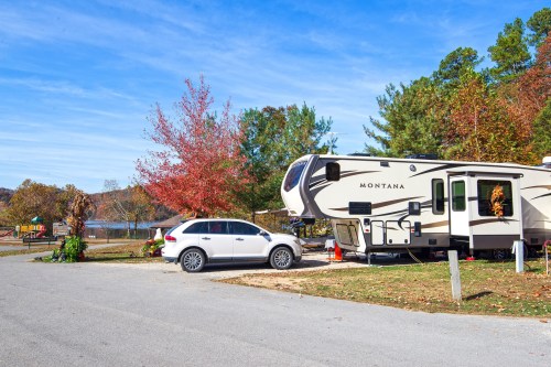 A parked RV with a car nearby, surrounded by trees with autumn foliage and a lake in the background.