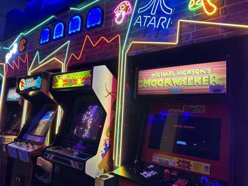 A row of colorful arcade machines with neon lights, featuring classic games like Pac-Man and Michael Jackson's Moonwalker.