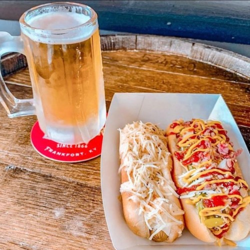 A frosty beer mug beside a plate of two hot dogs topped with sauerkraut, mustard, and ketchup on a wooden table.