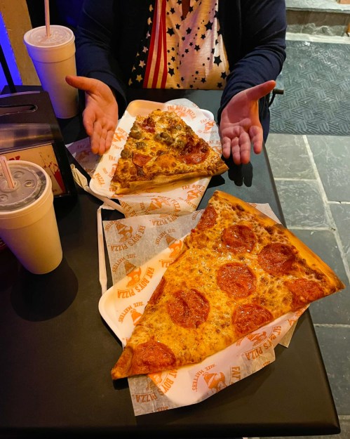 Two large slices of pepperoni pizza on paper plates, with a person’s hands gesturing beside them.