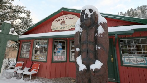 A wooden bear statue stands in front of a red candy shop with snow on the roof and chairs outside.