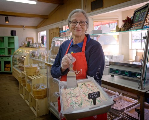 A smiling woman in an apron holds a tray of candy cane treats in a cozy shop filled with sweets.