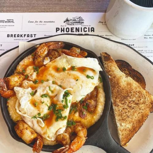 A skillet dish featuring shrimp, grits, a fried egg, and toast, served on a restaurant table with a coffee cup.