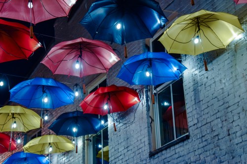 Colorful umbrellas in red, blue, and yellow hang above a street, illuminated by warm lights against a brick wall.