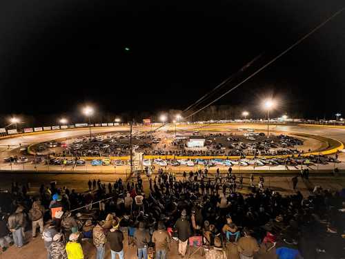 Aerial view of a busy racetrack at night, filled with cars and a large crowd of spectators. Bright lights illuminate the scene.