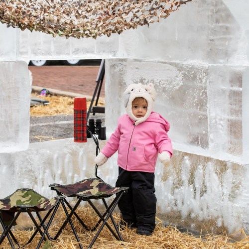 A toddler in a pink jacket and animal hat stands beside folding chairs in a winter setting.