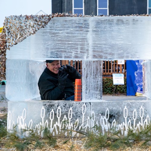 A person smiles while sitting inside a large ice sculpture, surrounded by ice blocks and a wooden backdrop.