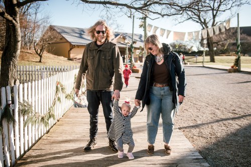 A family walks together on a path, holding hands, with a child in a checkered outfit and parents smiling.