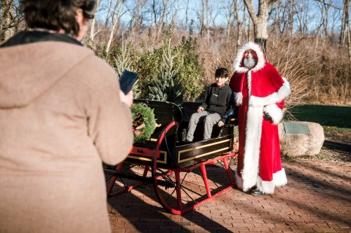 A child sits in a festive sleigh with Santa Claus, while a person takes a photo from behind. Trees in the background.