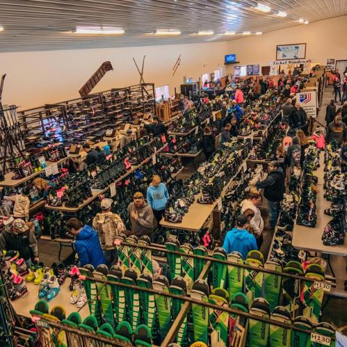 A busy indoor shoe store with rows of shoes and many customers browsing and shopping.