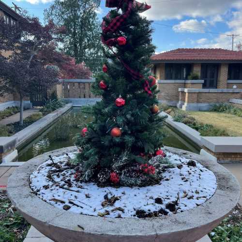 A small Christmas tree decorated with ornaments sits in a snowy planter, surrounded by greenery and a pond.